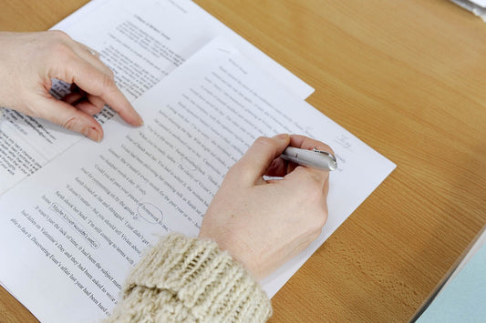 A close up of someone manually editing a two-page, printed document. Words in the document are crossed out, underlined and circled in pen. Only the hands and wrists of the editor are visible as they lean over the document, a pen in their right hand.