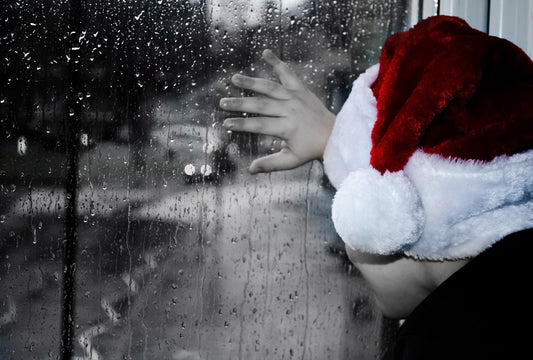 An image of a young child wearing a santa hat and staring out of a rain-washed window at a blurred street. Only the santa hat is coloured, in deep red and white. The rest of the image is greyscale.